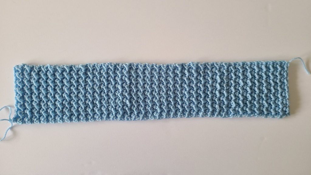Rectangular crocheted blue headband panel shown flat, displaying the textured stitch pattern before sewing the twist.