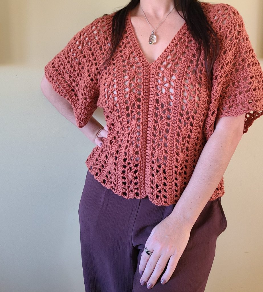 A person stands with one hand on their hip, wearing a rust-colored crochet lace top made with chunky yarn. The top has a V-neck, airy openwork patterns, wide sleeves, and a slightly scalloped hem, paired with loose mauve trousers.