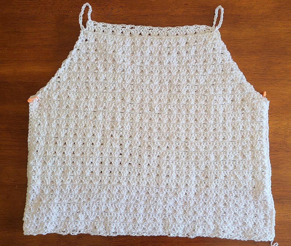 Flat lay of a crocheted top panels worked in a textured stitch pattern, shown after shaping for the armholes and neckline, laid on a wooden surface with stitch markers on each side.