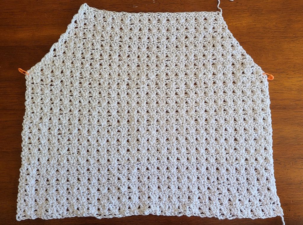Flat lay of a crocheted top panel worked in a textured stitch pattern, shown after shaping for the armholes and neckline, laid on a wooden surface with stitch markers on each side.