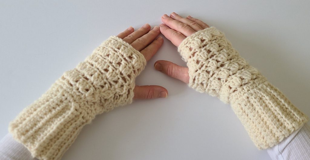 “Close-up of two hands wearing beige crochet fingerless gloves displayed on a flat white background, showing the texture and stitch details.”