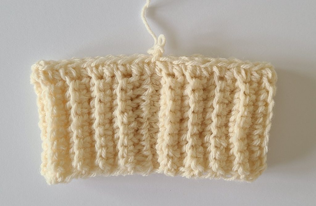 One beige half crochet leg warmer displayed on a flat white background, showing texture and stitch details, the bottom ribbing and the first row for the body section.