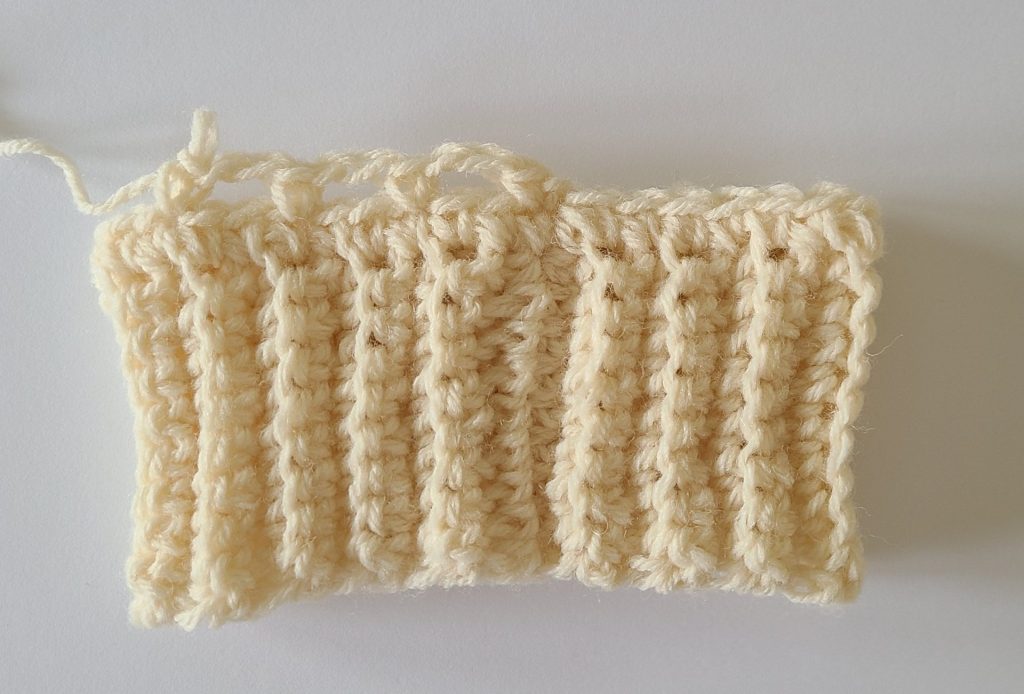 One beige half crochet leg warmer displayed on a flat white background, showing texture and stitch details, the bottom ribbing and and how to start row 2.