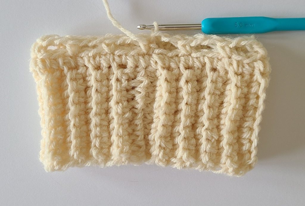 One beige half crochet leg warmer displayed on a flat white background, showing texture and stitch details, the bottom ribbing and and how to finish row 2.