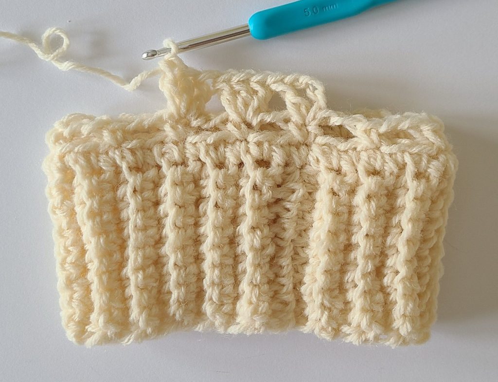 One beige half crochet leg warmer displayed on a flat white background, showing texture and stitch details, the bottom ribbing and and how to start row 3.