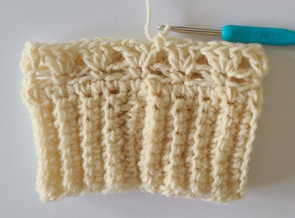 One beige half crochet leg warmer displayed on a flat white background, showing texture and stitch details, the bottom ribbing and and how to finish row 3.