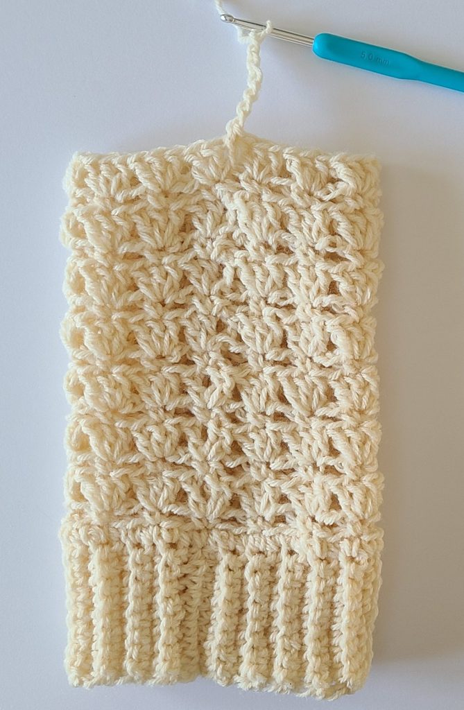 One beige crochet leg warmer displayed on a flat white background, showing texture and stitch details starting a top ribbing
