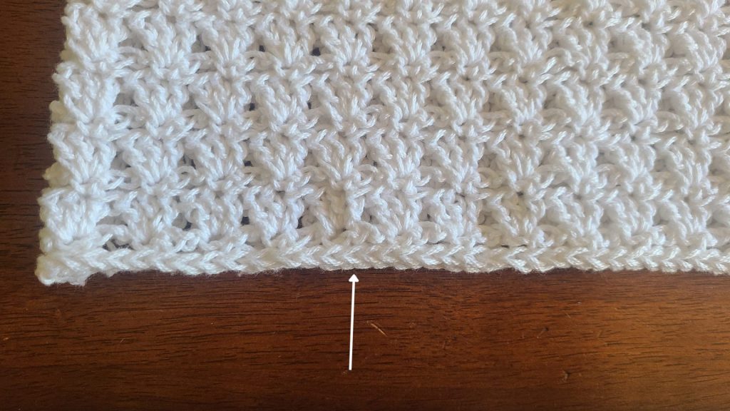 Flat lay of a bottom crocheted top panel worked in a textured stitch pattern, shown after shaping for the armholes and neckline, laid on a wooden surface with stitch markers on each side.