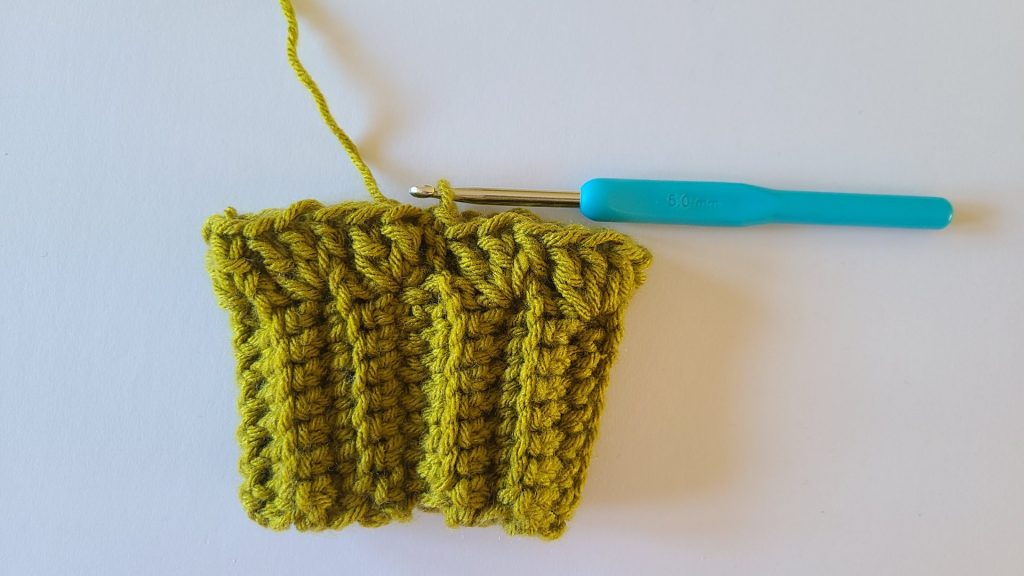 "Close-up of olive green crochet fingerless gloves, focusing on the ribbed texture. Row 1 is fully completed, showing all stitches clearly from start to finish, with each stitch and the overall pattern visible for reference."