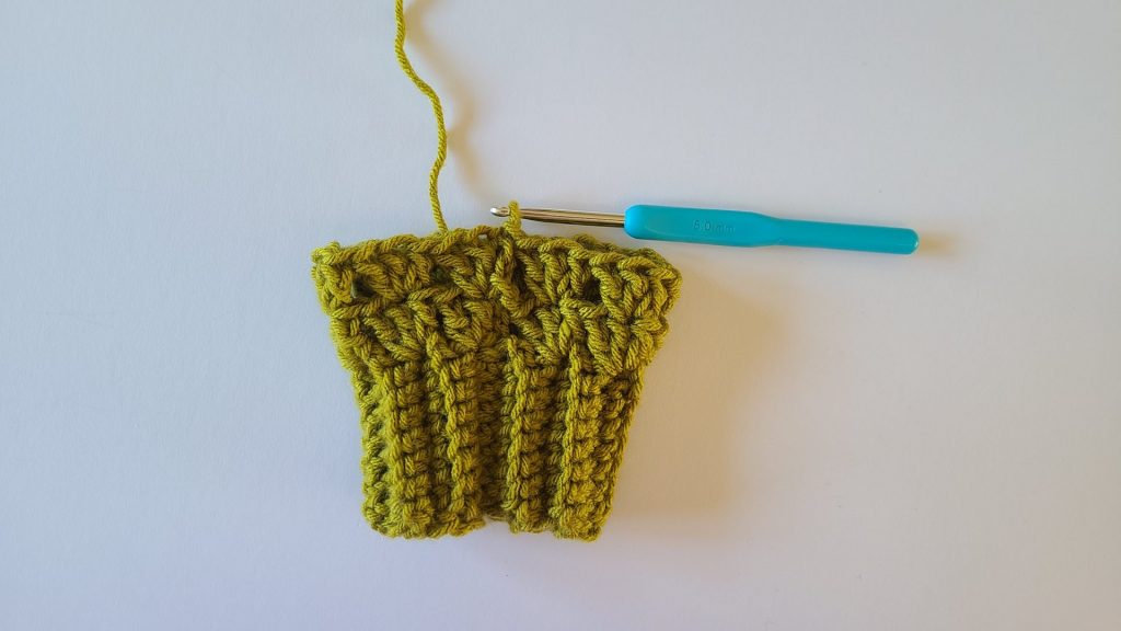 "Close-up of olive green crochet fingerless gloves, highlighting the textured ribbing. Row 2 is fully completed, showing all stitches clearly, the pattern in relief, and the overall structure of the glove for reference."