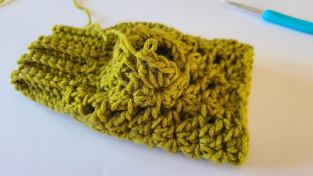"Close-up of an olive green crochet fingerless glove laid flat on a white background, showing the thumb border, stitch placement, and ribbed texture for construction reference."