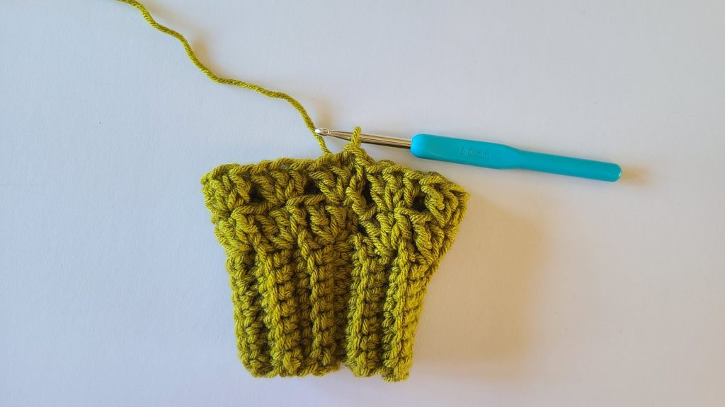 "Close-up of olive green crochet fingerless gloves showing textured ribbing and the completed second row, with stitches and pattern clearly visible."