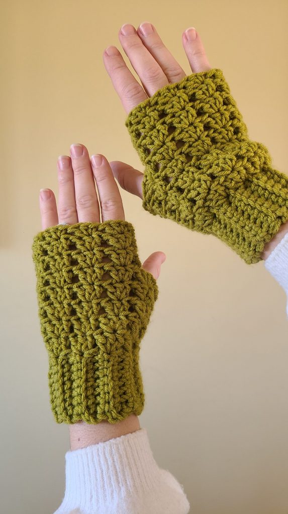 Hands wearing olive green crochet fingerless gloves with a textured, lacy stitch pattern against a neutral background.