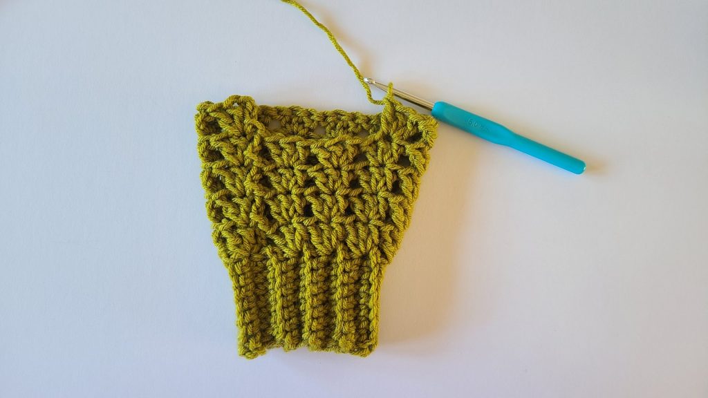 "Close-up of olive green crochet fingerless gloves, highlighting the textured ribbing. Row 5 is fully completed, showing all stitches clearly, the pattern in relief, and the overall structure of the gloves for reference."