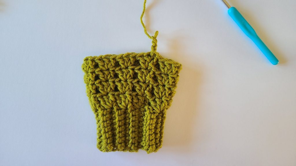 "Olive green crochet fingerless gloves laid flat on a white background, showing the fully completed row 4. The ribbed texture and individual stitches are clearly visible, highlighting the pattern, structure, and overall design of the gloves."