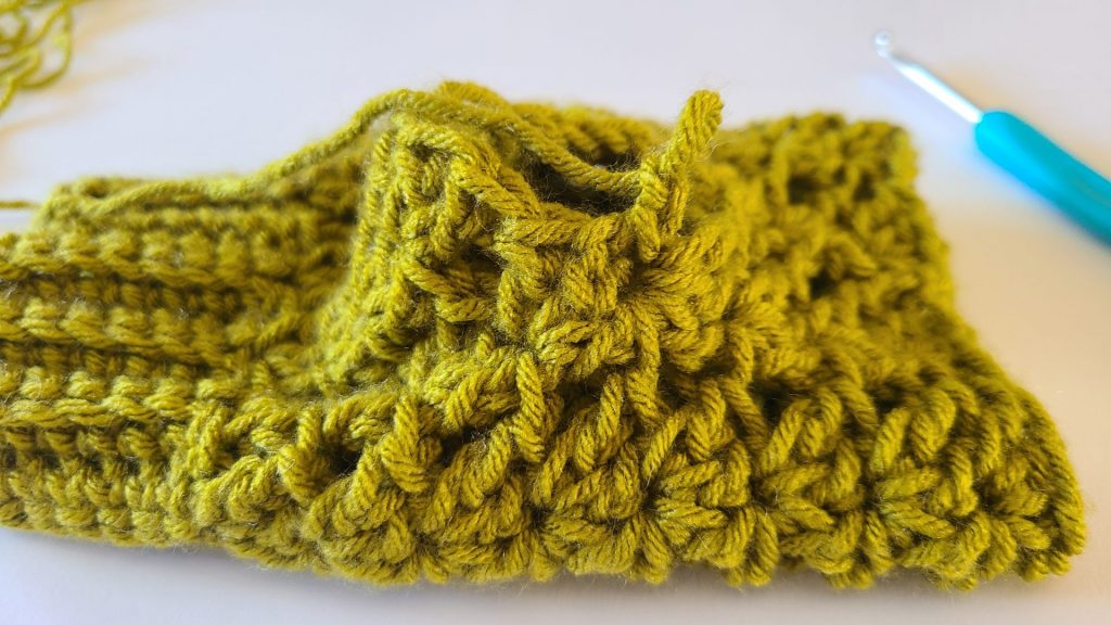 "Olive green crochet fingerless glove laid flat on a white background, highlighting the thumb border, ribbed texture, and stitch details around the thumb opening."